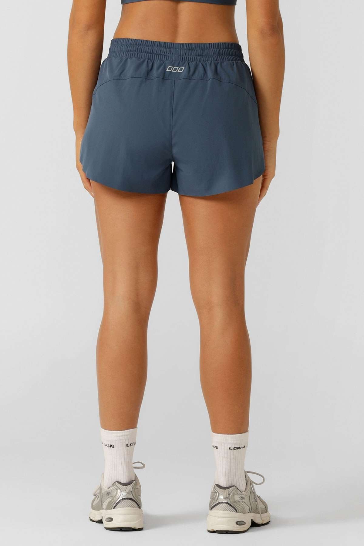 A person stands facing away, wearing a dark sports bra and blue Lorna Jane Lightspeed Phone Pocket Run Shorts with sweat-wicking fabric, plus white socks and gray sneakers, set against a plain background.