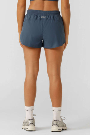 A person stands facing away, wearing a dark sports bra and blue Lorna Jane Lightspeed Phone Pocket Run Shorts with sweat-wicking fabric, plus white socks and gray sneakers, set against a plain background.