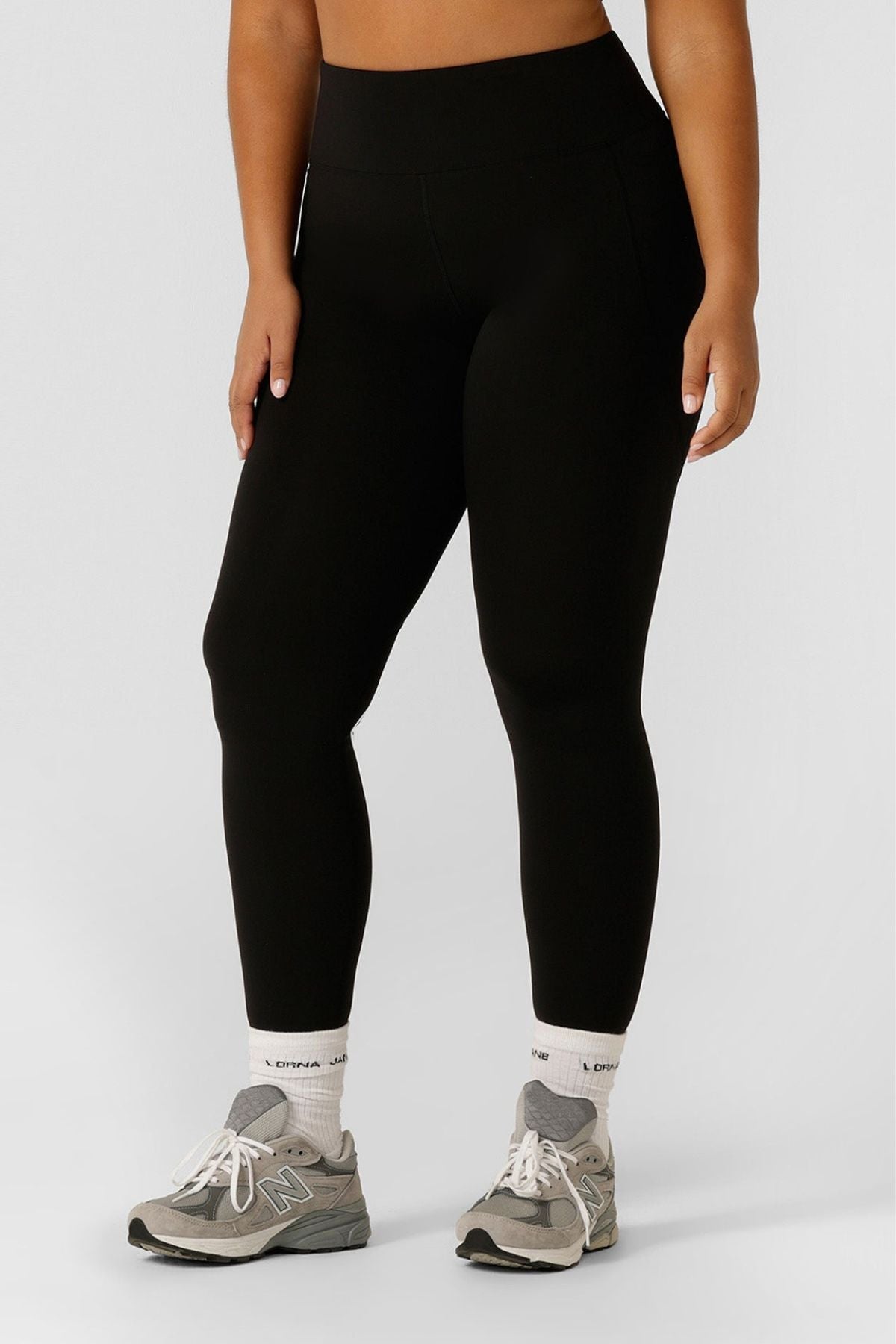 A person wears Lorna Jane Amy Thermal Phone Pocket Ankle Biter Leggings, white crew socks, and gray New Balance sneakers against a plain light background. Only their lower torso and legs are visible.