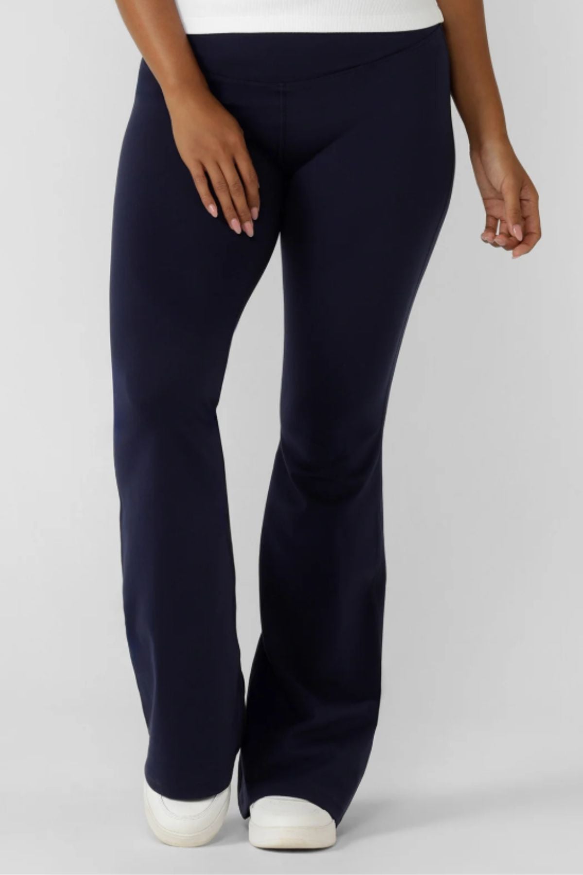A person wearing Lorna Jane Lotus Flared Legging in navy blue and white sneakers stands against a plain light background, showing only the lower torso, legs, and one arm.