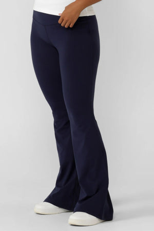 Model wears the Lorna Jane Lotus Flared Legging in navy, featuring a high-rise waistband and flared legs, paired with white sneakers; shown from the waist down against a plain background.