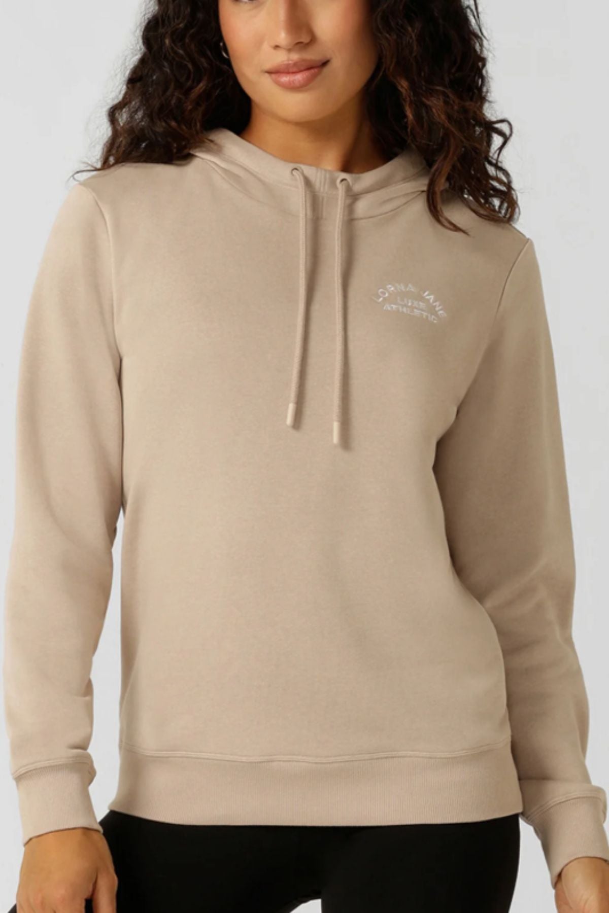 A woman wears the Lorna Jane Lotus Hoodie, a lightweight beige cotton-blend hoodie with drawstrings and a small white logo on the chest, standing against a plain light background.