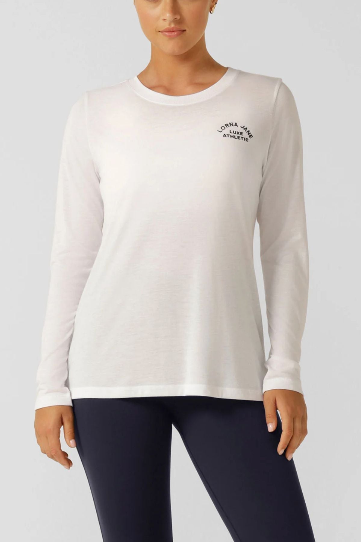 Woman in the Lorna Jane Lotus Long Sleeve Top featuring "Lorna Jane Athlete" on the chest, paired with black leggings, standing against a light gray background.