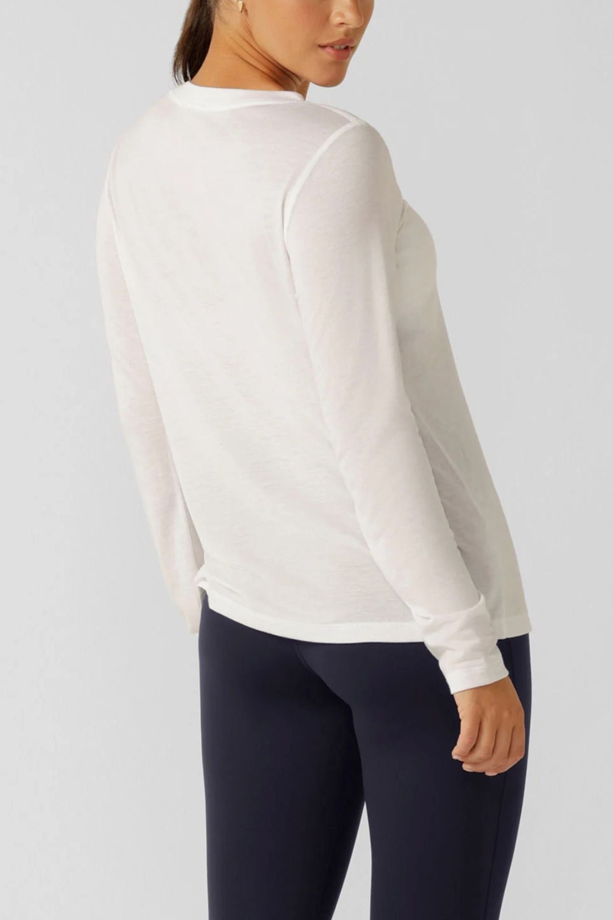 A woman wearing the Lorna Jane Lotus Long Sleeve Top with dark pants stands facing away, looking over her shoulder against a plain light background—showcasing chic activewear style.