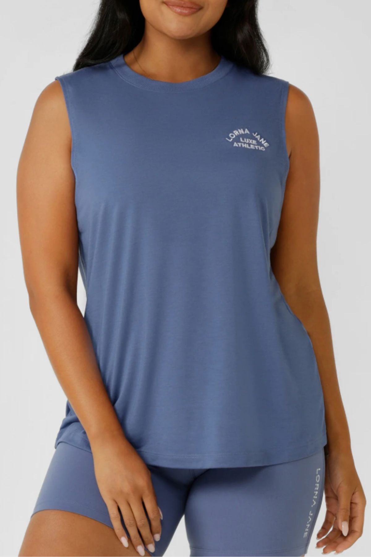 A woman wears the Lorna Jane Lotus Muscle Tank, a lightweight, breathable top perfect for layering, as she poses with one hand by her side.