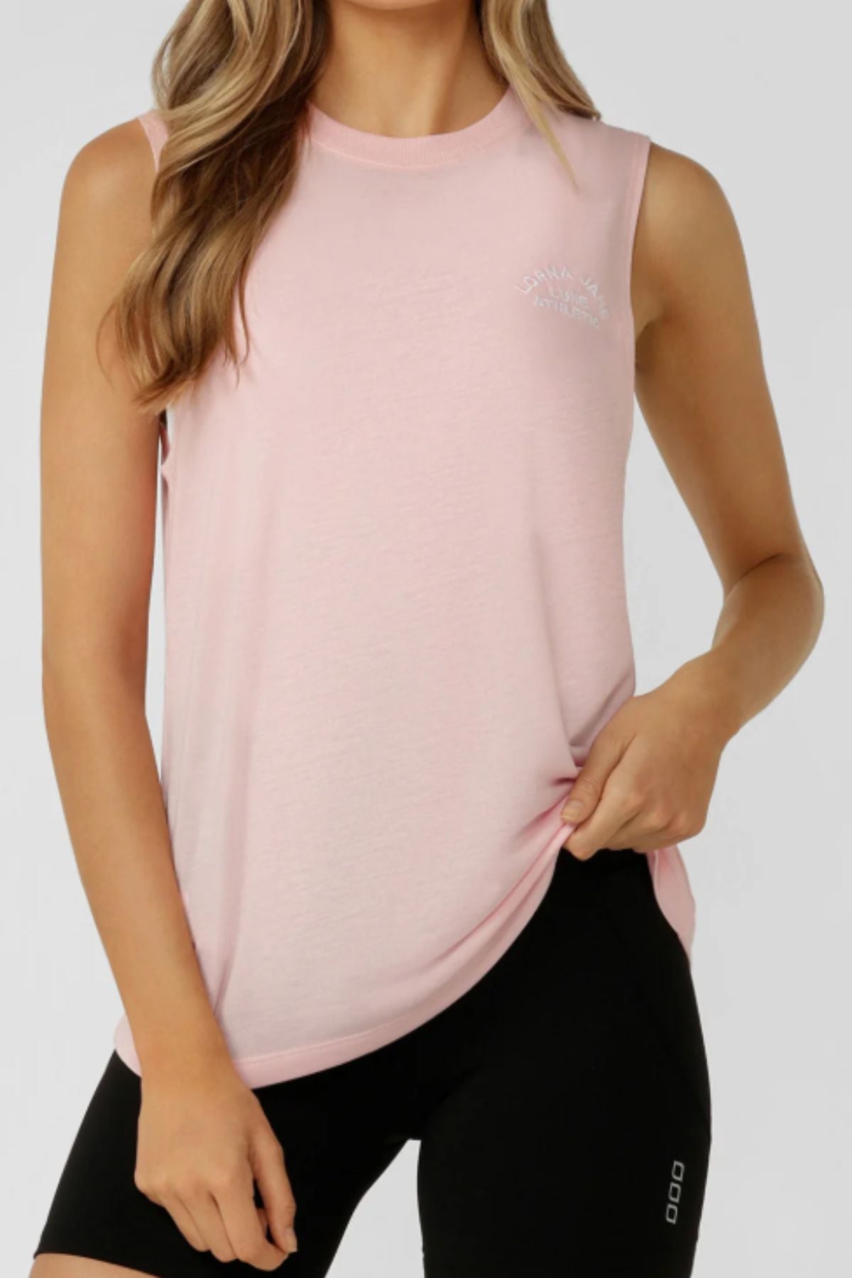 A woman wears the Lorna Jane Lotus Muscle Tank in light pink with black shorts, standing against a plain background and holding the hem of her shirt with one hand.
