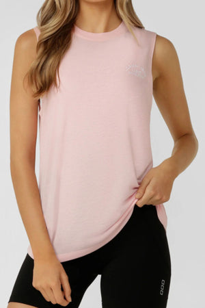 A woman wears the Lorna Jane Lotus Muscle Tank in light pink with black shorts, standing against a plain background and holding the hem of her shirt with one hand.