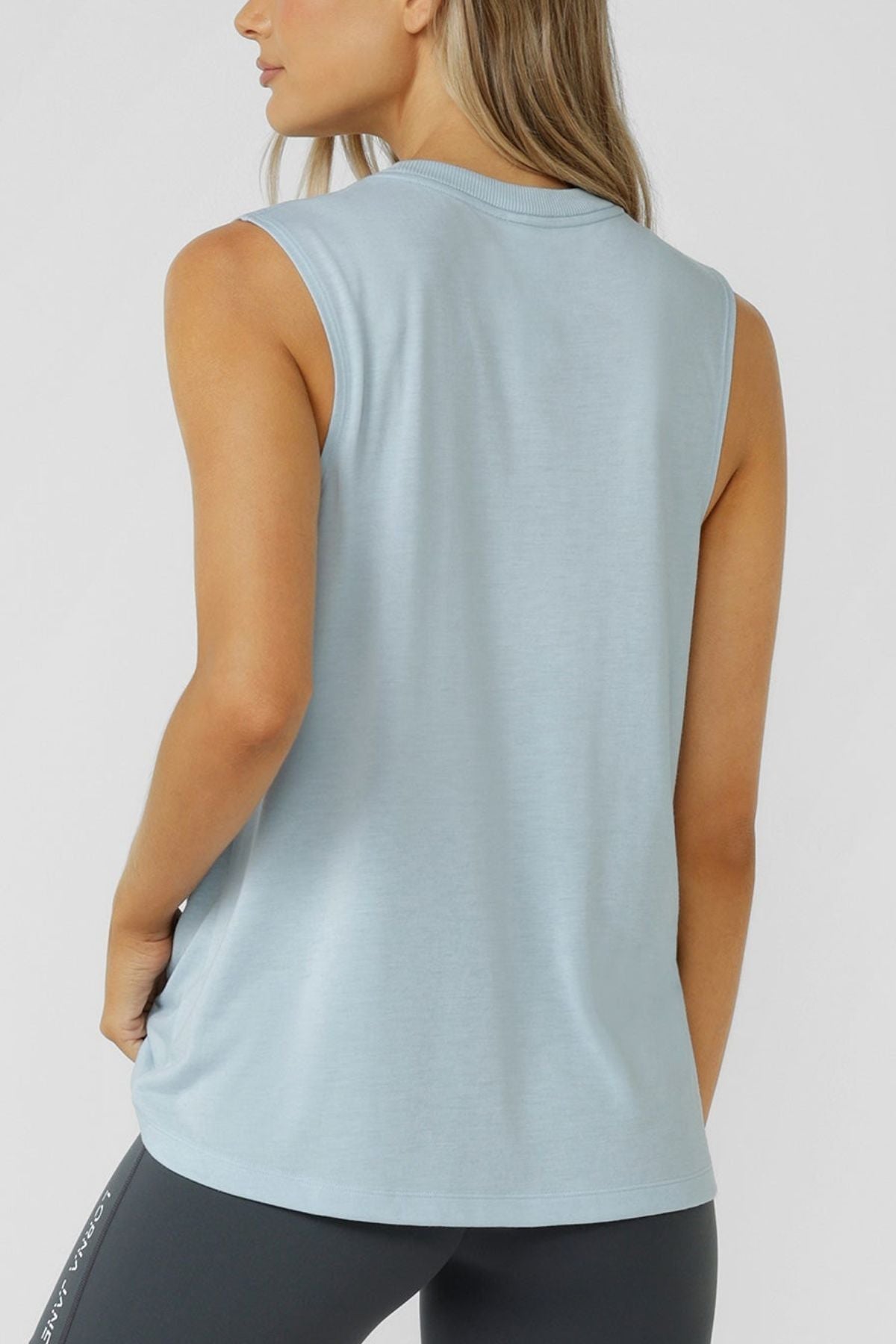 A person with long, light brown hair is shown from the back against a plain white background, wearing a sleeveless light blue Lorna Jane Lotus Muscle Tank paired with dark leggings.
