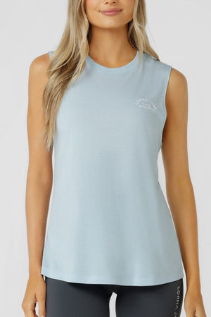 A woman with long blonde hair and a slight smile stands against a plain background, dressed in dark leggings and the Lorna Jane Lotus Muscle Tank in light blue. This breathable tank features a small white logo on the left chest, making it ideal for lightweight layering.
