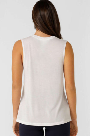 A woman with long brown hair is seen from the back, wearing the Lorna Jane Lotus Muscle Tank—a sleeveless white top—paired with dark pants, set against a plain light gray background.