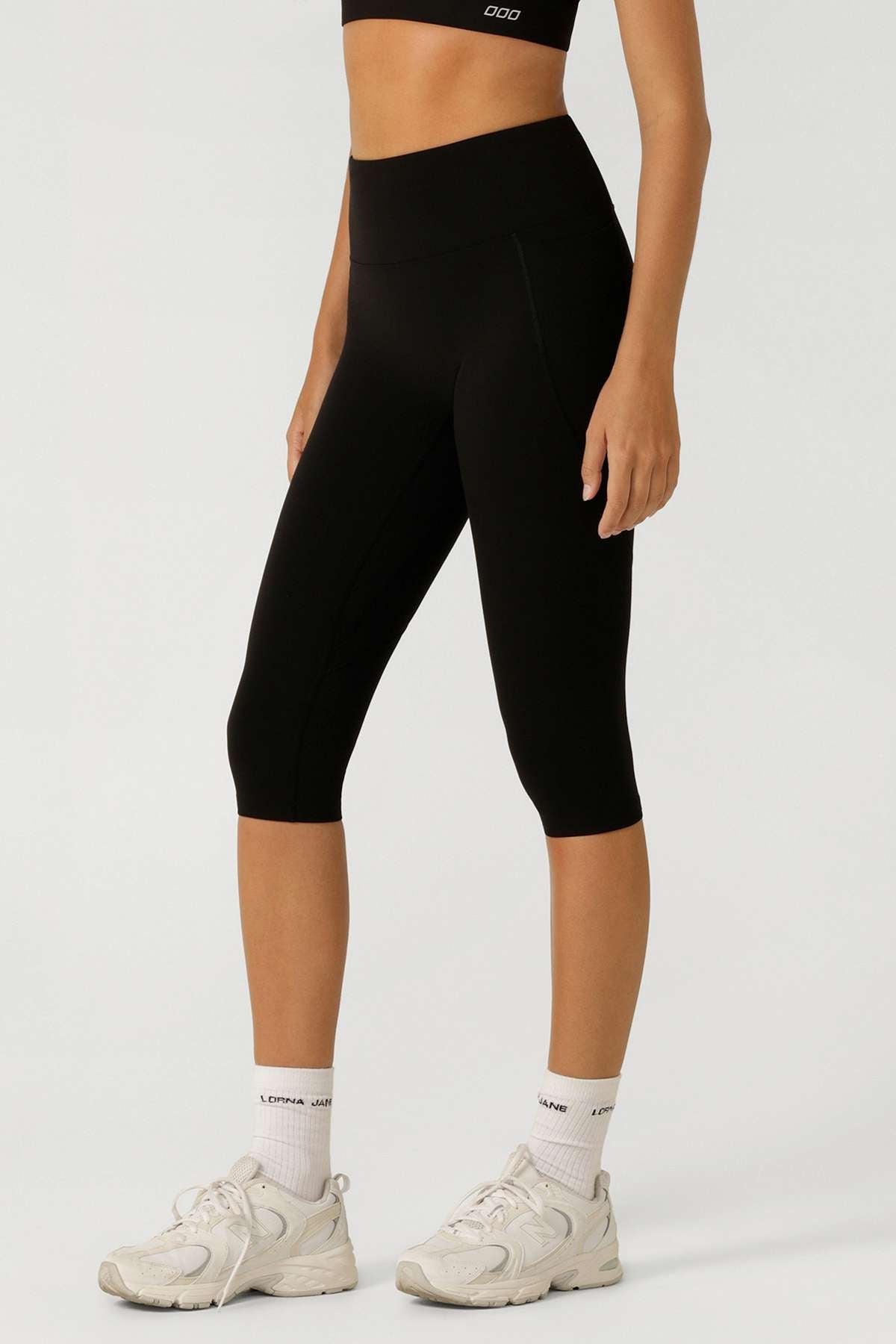 A person in a black sports bra and Lorna Jane Lotus No Chafe 7/8 Leggings stands against a plain light background. These high-rise, moisture-wicking leggings are ideal for active days, paired with white socks and athletic shoes.