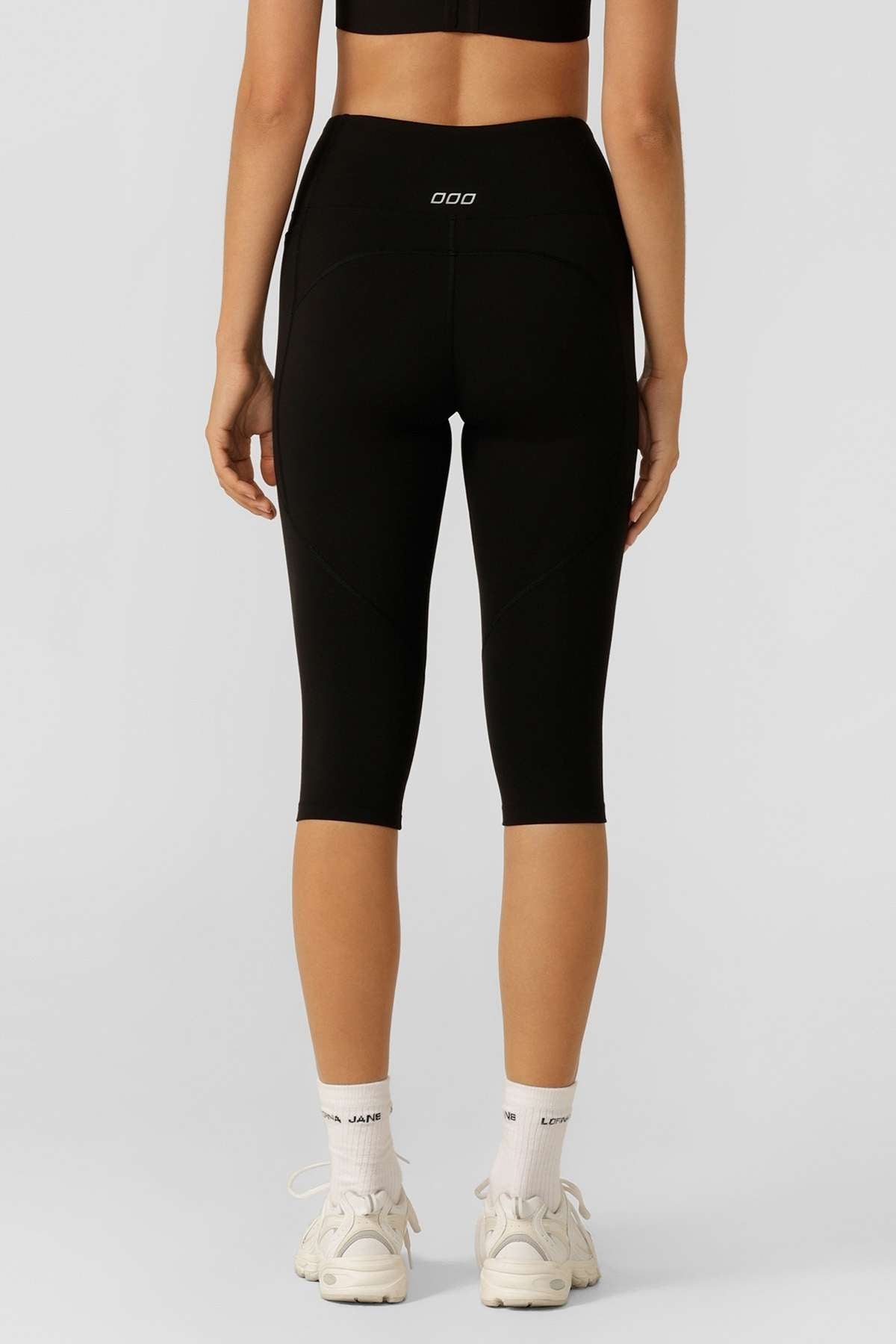 A person shown from the back, wearing black Lorna Jane Lotus No Chafe 7/8 Leggings, white crew socks, and white sneakers stands against a plain light gray background.