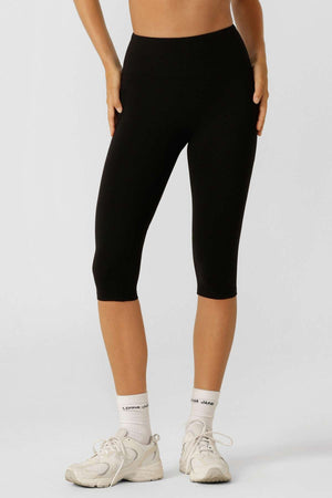 A person wears black Lorna Jane Lotus No Chafe 7/8 Leggings, white socks, and athletic shoes, standing against a plain light background. The photo is cropped from midriff to feet.