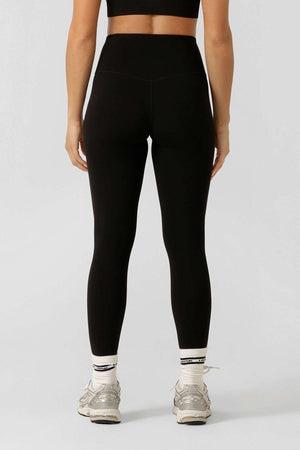 A woman faces away, wearing a black sports bra and Lorna Jane Lotus No Chafe Ankle Biter Leggings made from 4-way stretch, moisture-wicking fabric, plus white socks and gray athletic shoes against a light gray background.