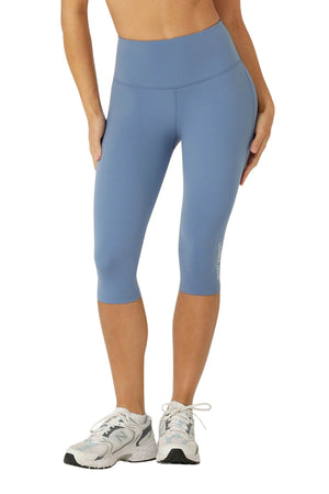 A person shown from mid-torso to feet wears Lorna Jane Lotus No Chafe Phone Pocket 3/4 Leggings in blue and white athletic sneakers against a white background. These leggings feature moisture-wicking fabric for workout comfort.