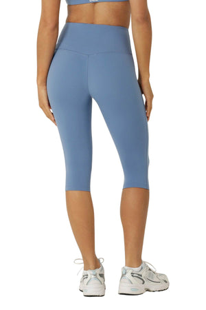 Woman wearing light blue Lorna Jane Lotus No Chafe Phone Pocket 3/4 Leggings and white sneakers, standing with her back to the camera against a white background.