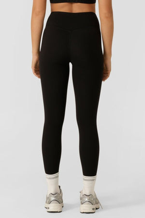 A person shown from the back against a plain light gray background wears black Lorna Jane Lotus No Chafe Phone Pocket Ankle Biter Leggings, white socks, and white athletic shoes.