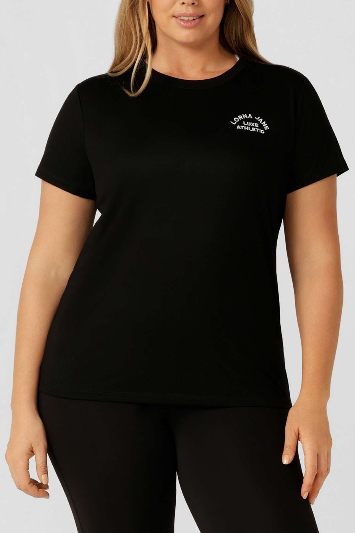A woman stands against a neutral background wearing the Lorna Jane Lotus T-Shirt, featuring white "Lorna Jane Athletic" text on the left chest—ideal for an active lifestyle.