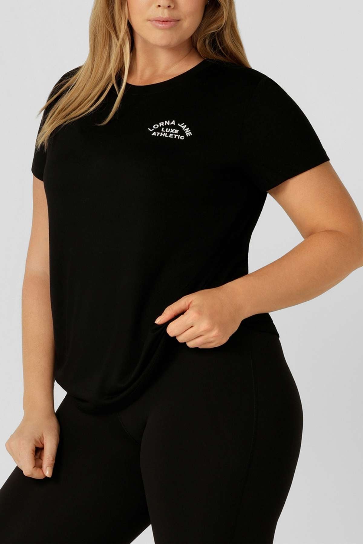 A woman stands in a plain light gray background, wearing the Lorna Jane Lotus T-Shirt with an embroidered logo on the left chest and matching black leggings, holding the hem. Ideal outfit for active lifestyles.