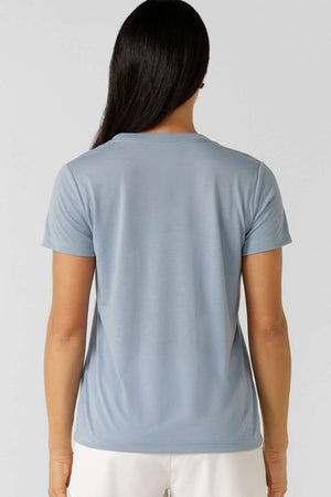 A person with long dark hair is seen from behind wearing a light blue Lorna Jane Lotus T-Shirt, featuring 2-way stretch, against a plain light background.