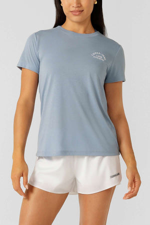 A woman stands against a plain light gray background wearing a light blue Lorna Jane Lotus T-Shirt with white chest text and white athletic shorts, styled for an active lifestyle.