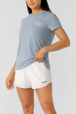 A woman in a light blue Lorna Jane Lotus T-Shirt featuring 2-way stretch and white shorts stands relaxed against a light gray background, showcasing an outfit ideal for an active lifestyle.