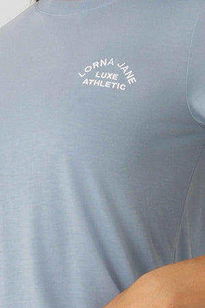 A close-up of the light blue Lorna Jane Lotus T-Shirt shows "Lorna Jane Luxe Athletic" in white text on the chest. The 2-way stretch fabric ensures comfort for an active lifestyle. Part of an arm is visible at the bottom right.
