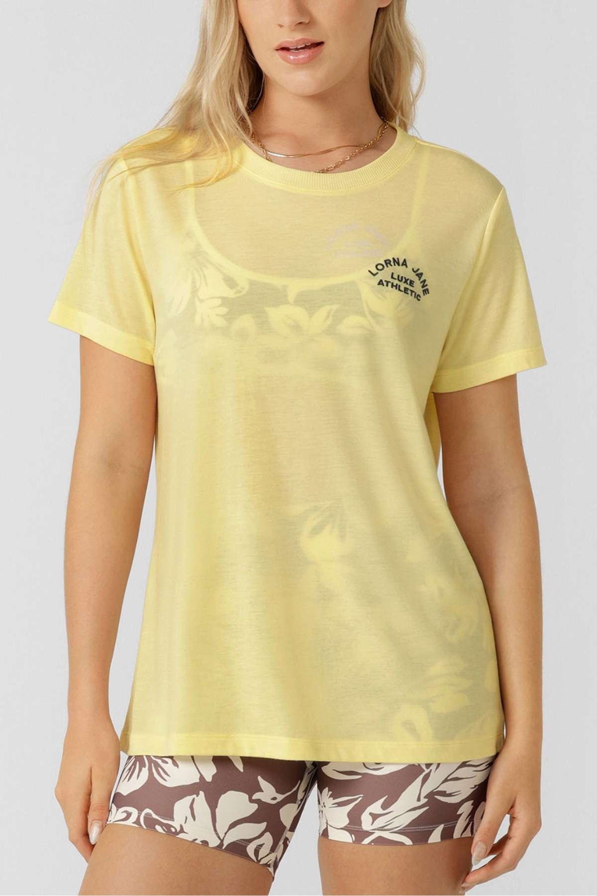 A woman with long blonde hair and a light necklace wears the Lorna Jane Lotus T-Shirt in light yellow with subtle floral details and text on the chest, paired with brown bike shorts with white florals for an active, stylish look.