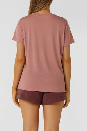 A woman with long, loose hair stands with her back to the camera wearing a mauve Lorna Jane Lotus T-Shirt and dark shorts, ideal for an active lifestyle. The scene features a plain, light-colored background.