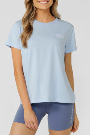 Smiling against a plain backdrop, a woman in the Lorna Jane Lotus T-Shirt and high-waisted blue biker shorts embodies an active lifestyle.