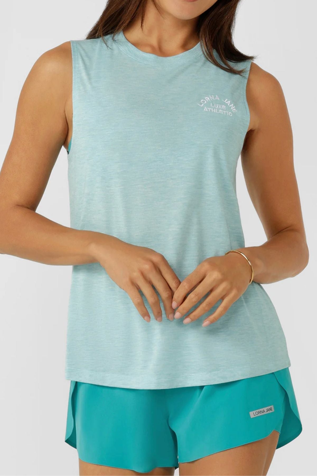 A woman wears the Lorna Jane Lotus Muscle Tank, styled in a relaxed long line fit in light blue with teal shorts. The top features a small white logo on the upper left chest as she stands with hands gently clasped at her waist.