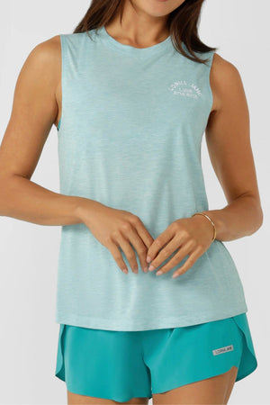 A woman wears the Lorna Jane Lotus Muscle Tank, styled in a relaxed long line fit in light blue with teal shorts. The top features a small white logo on the upper left chest as she stands with hands gently clasped at her waist.
