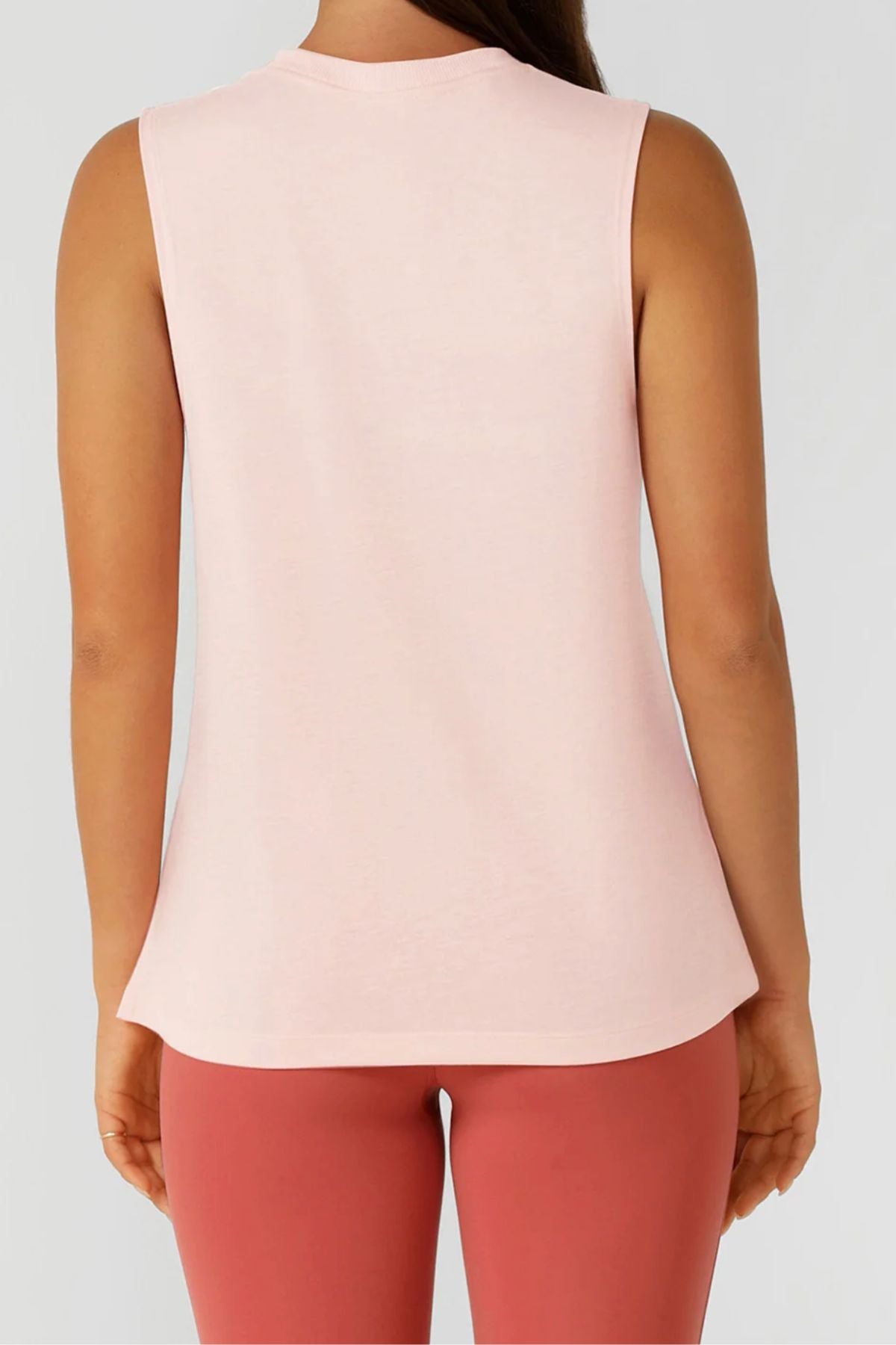 A person is seen from the back wearing a sleeveless, light pink Lorna Jane Lotus Muscle Tank with high-waisted, rose leggings against a plain light gray background.
