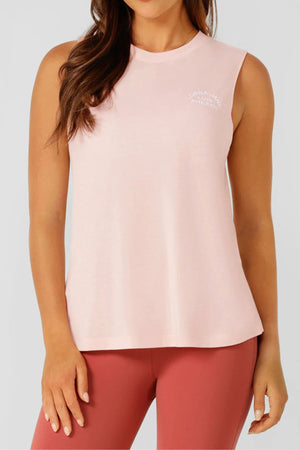 A person wearing the Lorna Jane Lotus Muscle Tank in light pink with white chest text, paired with high-waisted mauve leggings, stands against a plain light gray background.