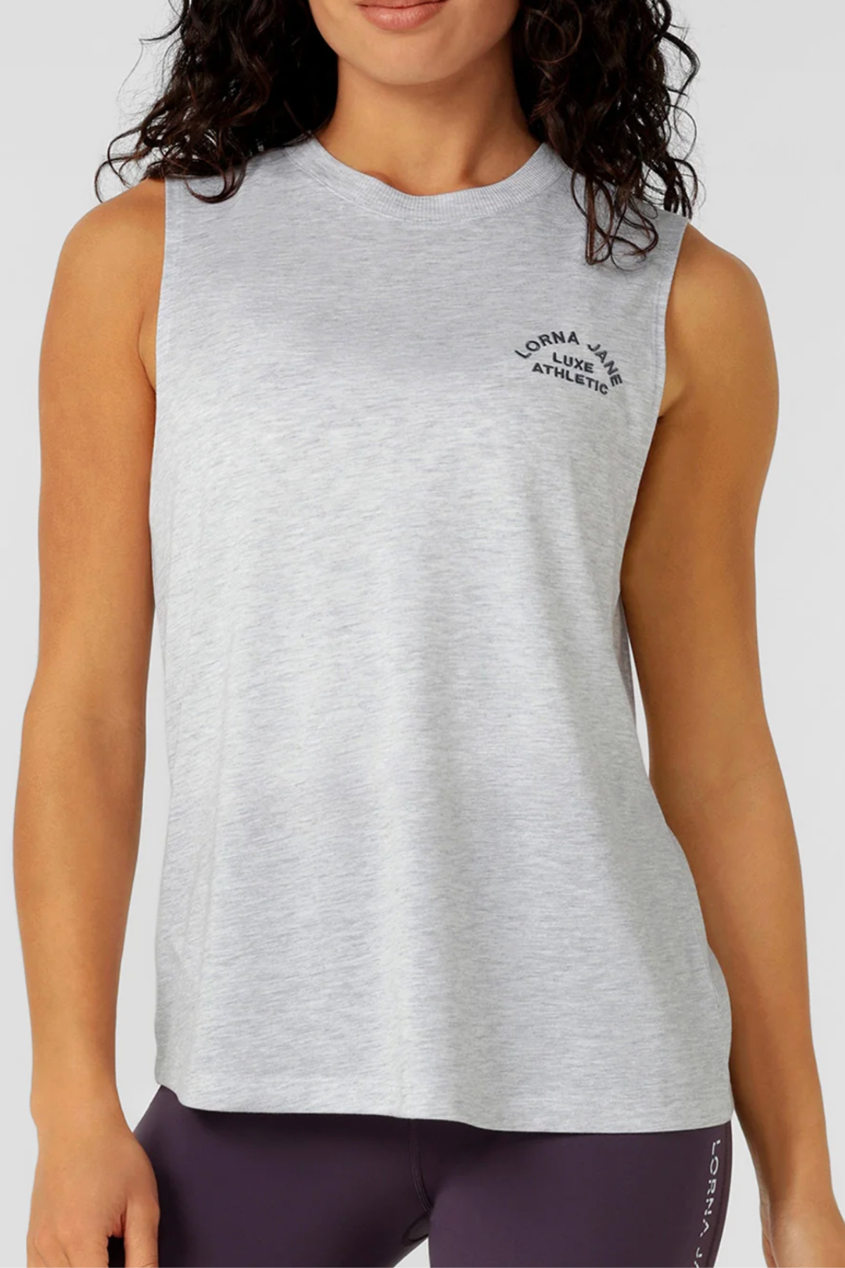 A woman wears a sleeveless, light gray Lorna Jane Lotus Muscle Tank with "Lorna Jane Athleisure" on the chest, paired with dark high-waisted leggings. The lightweight tank is cropped at the shoulders and thighs.