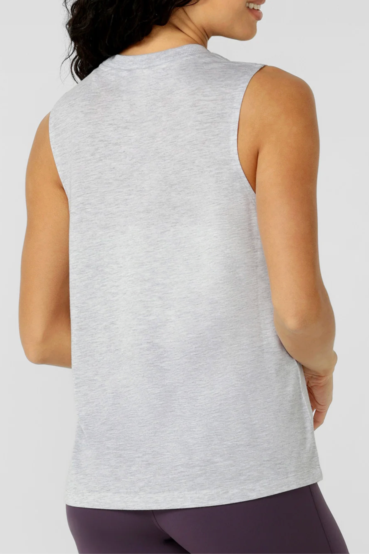 A woman with medium skin and wavy dark hair models the back of the Lorna Jane Lotus Muscle Tank in light gray, paired with dark purple leggings. The plain background highlights its lightweight, breathable natural blend fabric.