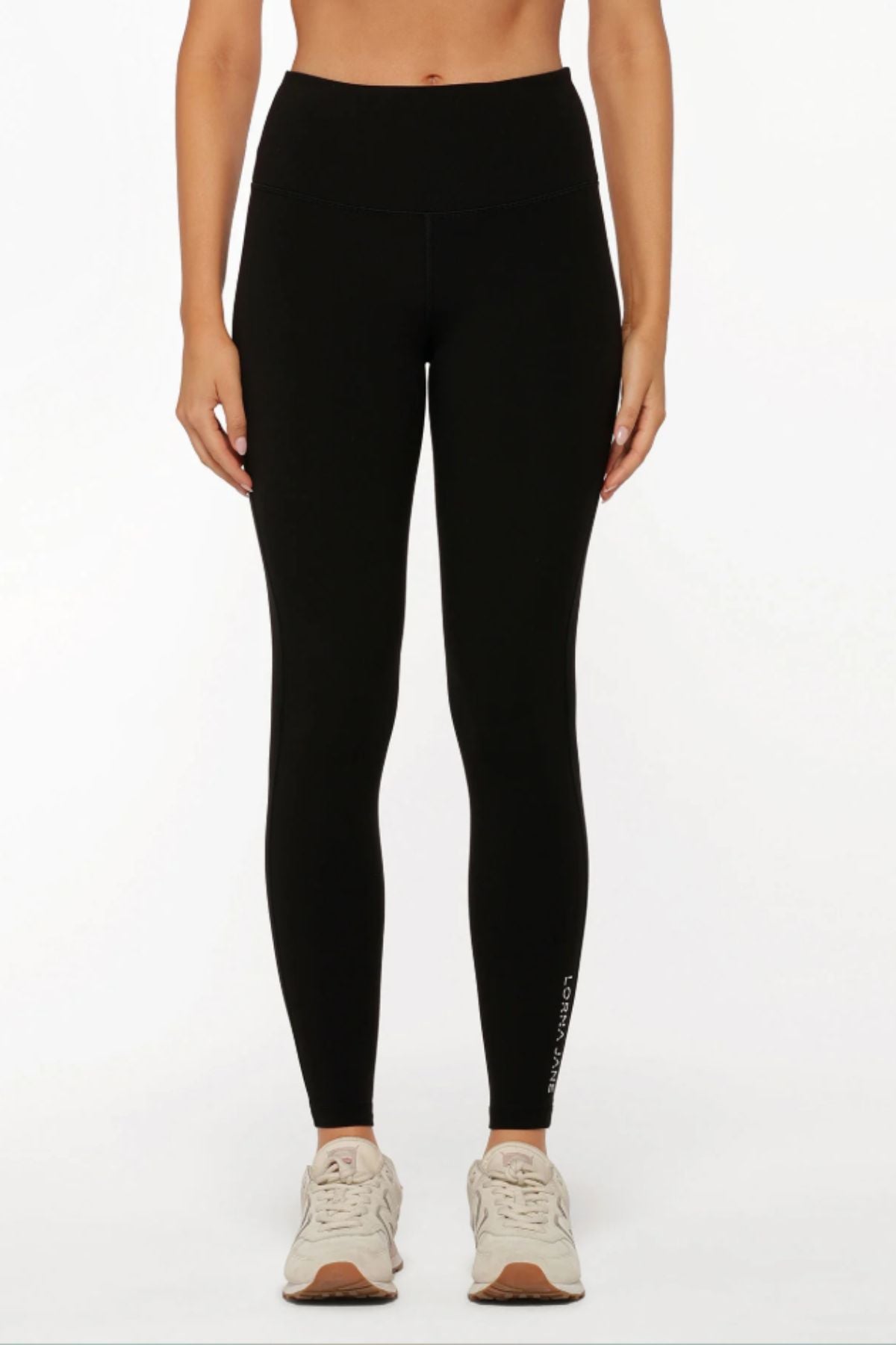 A person stands against a plain white background wearing Lorna Jane Lotus No Chafe Phone Pocket Full Length Leggings in black with a small white logo near the left ankle and beige athletic shoes.