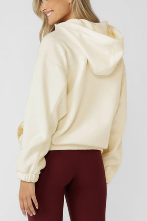 A woman with long blonde hair is shown from the back, smiling in a Lorna Jane Nova Polar Fleece Quarter Zip Hoodie and high-waisted burgundy leggings, standing against a plain light background.