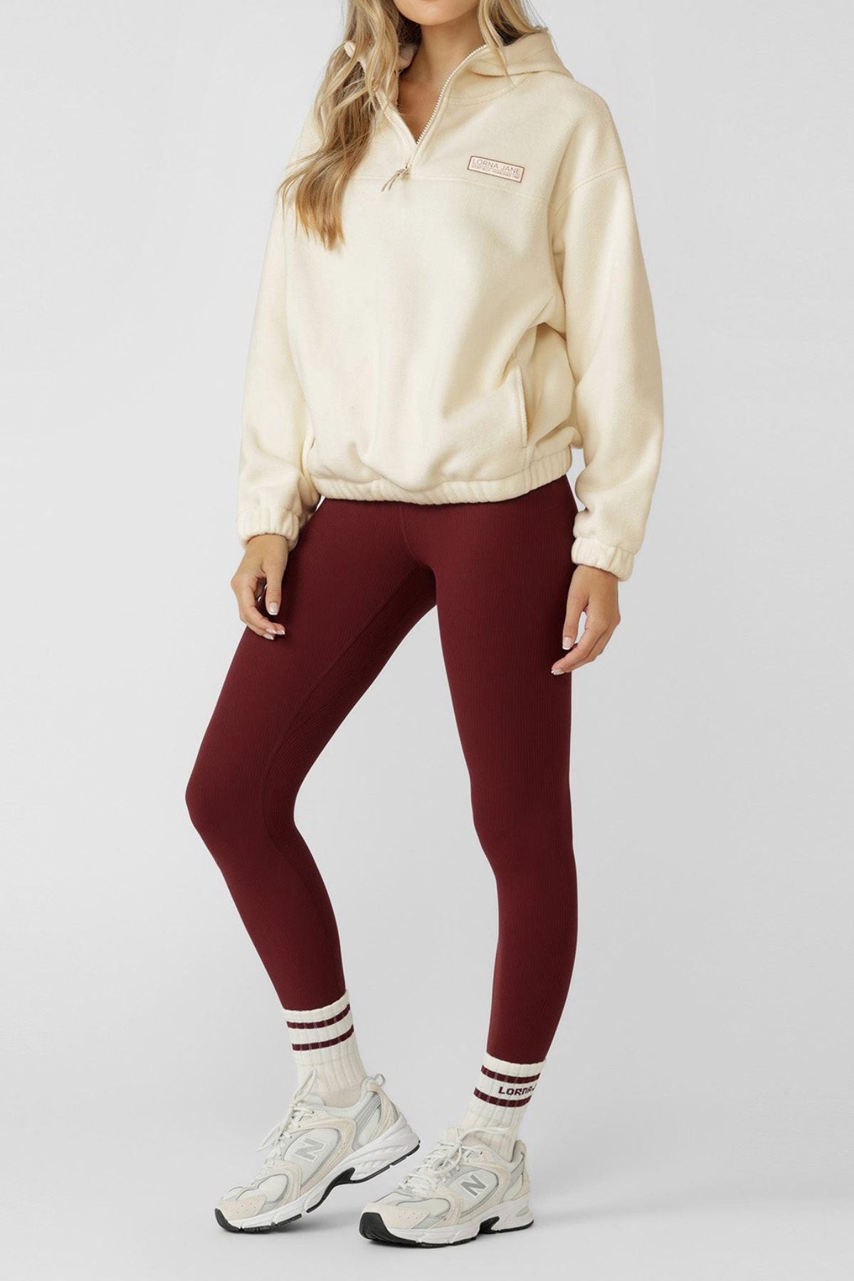 Woman wearing a cream Lorna Jane Nova Polar Fleece Quarter Zip Hoodie, burgundy leggings, white athletic shoes, and white socks with red stripes stands facing forward with hands relaxed by her sides against a plain light background.