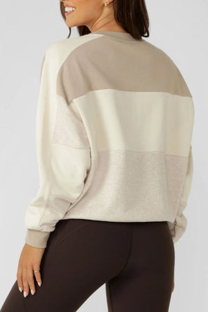 A woman with long brown hair is seen from the back, smiling slightly, wearing the Lorna Jane Lorna Luxe Panelled Sweat in beige and cream cotton-blend terry and dark brown leggings.