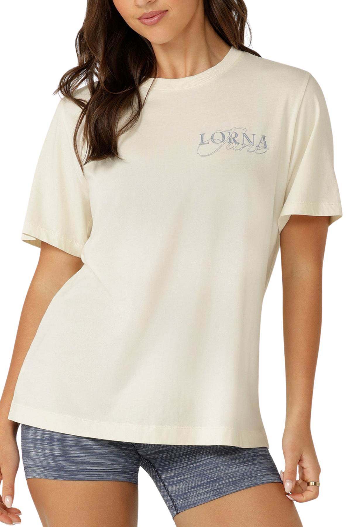A woman wears the Lorna Jane Paradise Postcard Washed Relaxed T-Shirt in premium soft cotton, featuring "Lorna" text on the chest, paired with blue-gray athletic shorts and posing with one hand on her hip.