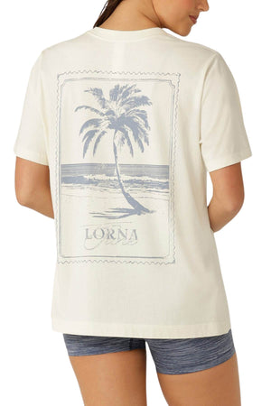 A woman wears the Lorna Jane Paradise Postcard Washed Relaxed T-Shirt in cream, featuring a large palm tree and "Lorna" on the back, paired with gray athletic shorts. The premium soft cotton tee is shown from behind.
