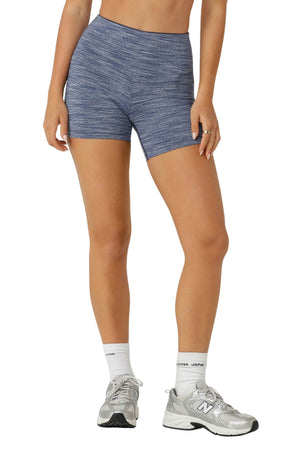 A person wears Lorna Jane Retro 12cm Bike Shorts in blue with a high-rise waistband, paired with white socks and silver running shoes. One hand rests on their thigh. The image is cropped from the waist down.
