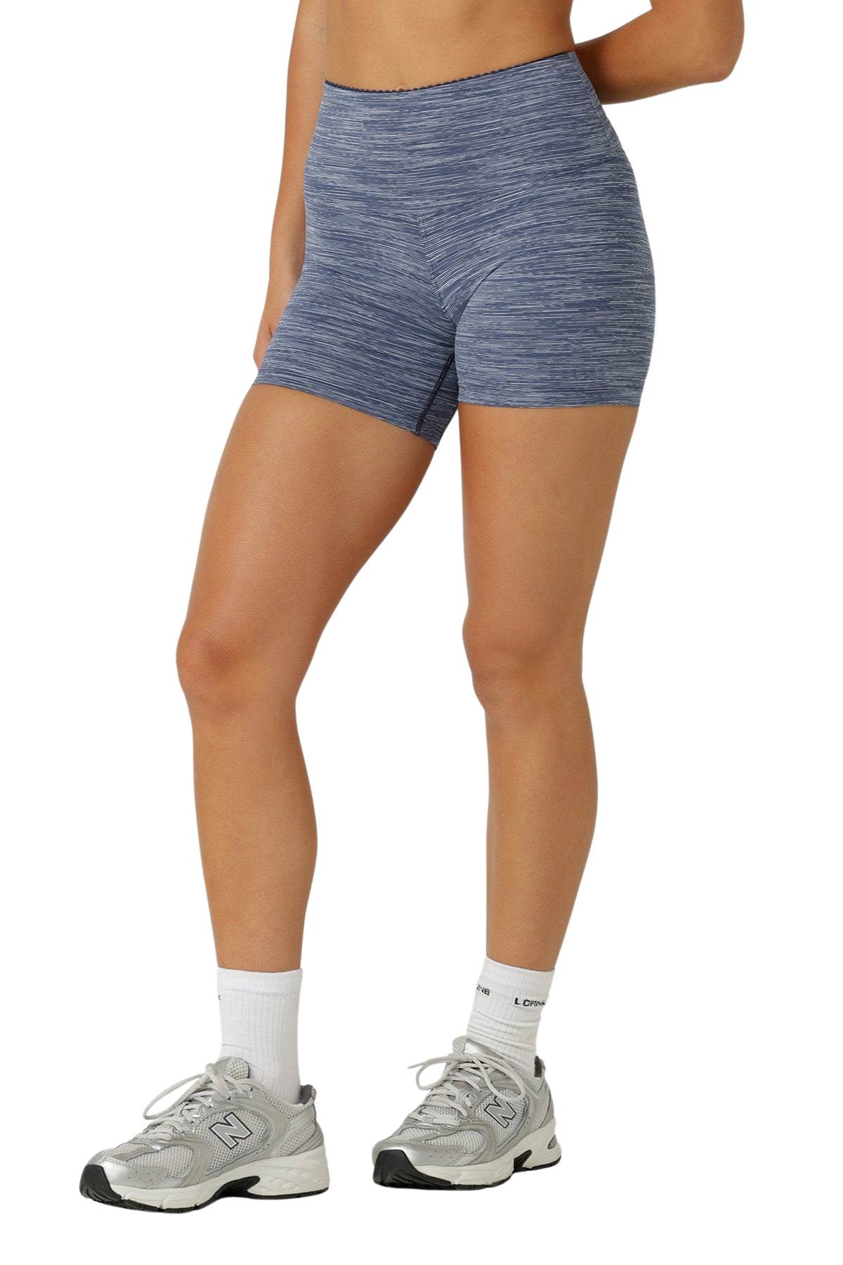A person wearing Lorna Jane Retro 12cm Bike Shorts in blue heather, paired with white crew socks and gray sneakers, stands with one arm cropped out and the other bent behind their back.