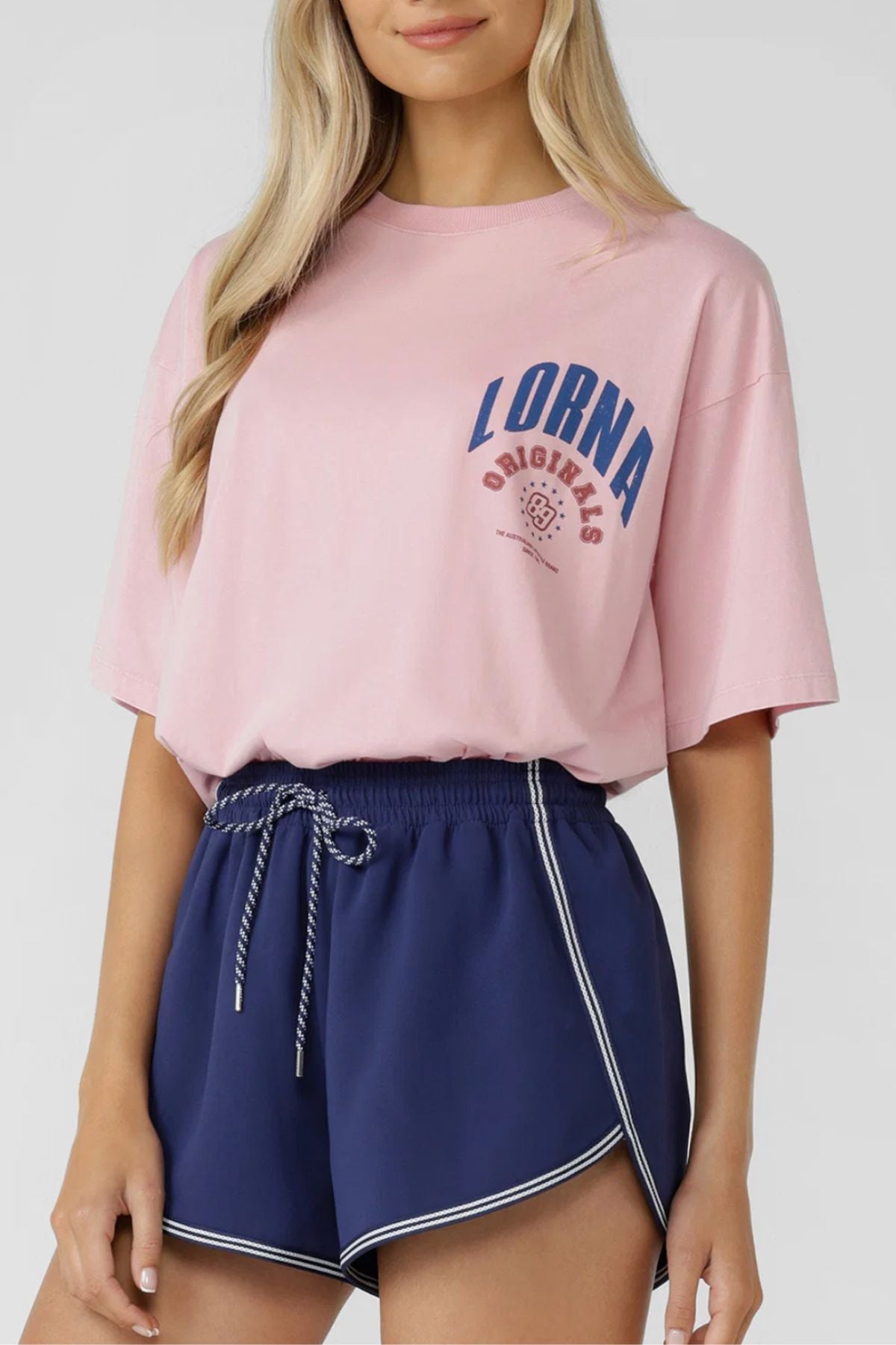 A woman wears the Lorna Jane Revival Washed Boyfriend T-Shirt in vintage pink with "LORNA ORIGINALS" blue curved lettering, paired with navy athletic shorts with white trim and a striped drawstring. 100% cotton for all-day comfort.