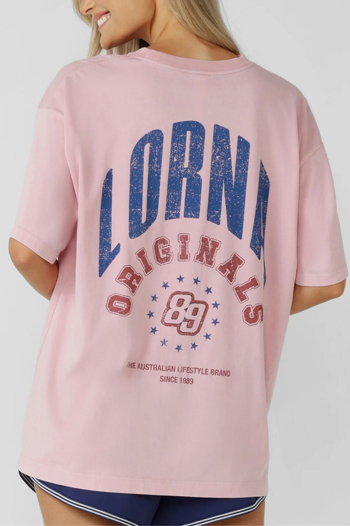 A woman with long blonde hair wears the Lorna Jane Revival Washed Boyfriend T-Shirt, featuring "Lorna Originals 89" in bold blue and red on the back. She is turned away, showing off the design, paired with navy blue shorts.