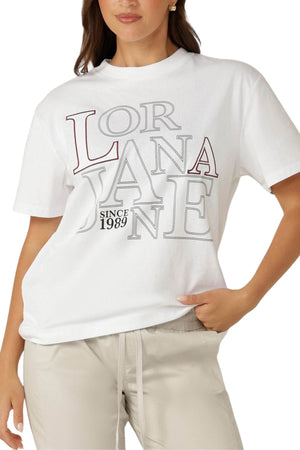 A woman wears the Lorna Jane Riviera T-Shirt, an oversized, premium cotton jersey tee with "LORNA JANE SINCE 1989" in bold letters. She pairs it with light beige pants; only her upper body is visible.