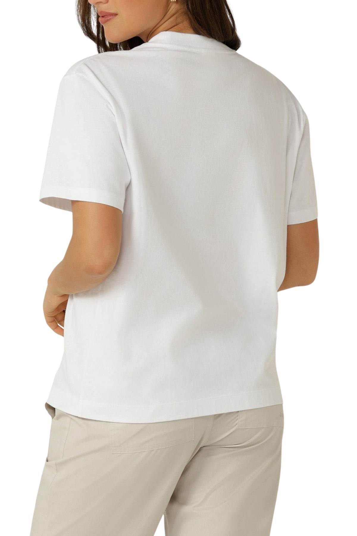 A person is seen from behind wearing a plain white Lorna Jane Riviera T-Shirt and light beige pants, with their left hand on their hip and head turned slightly left.