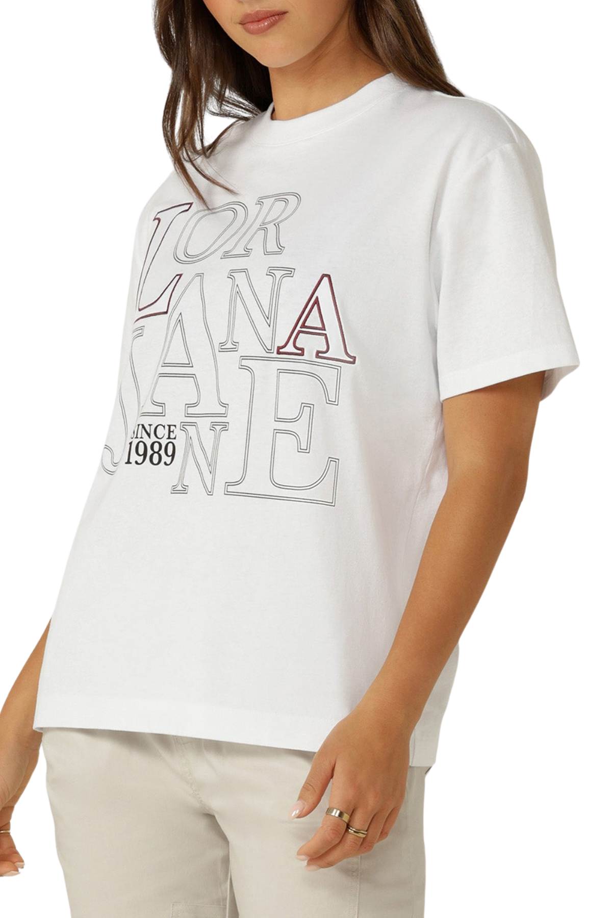 A woman wears a white, oversized Lorna Jane Riviera T-Shirt featuring large outlined lettering and "Since 1989" on the front, paired with light-colored pants. Only her upper body is visible.