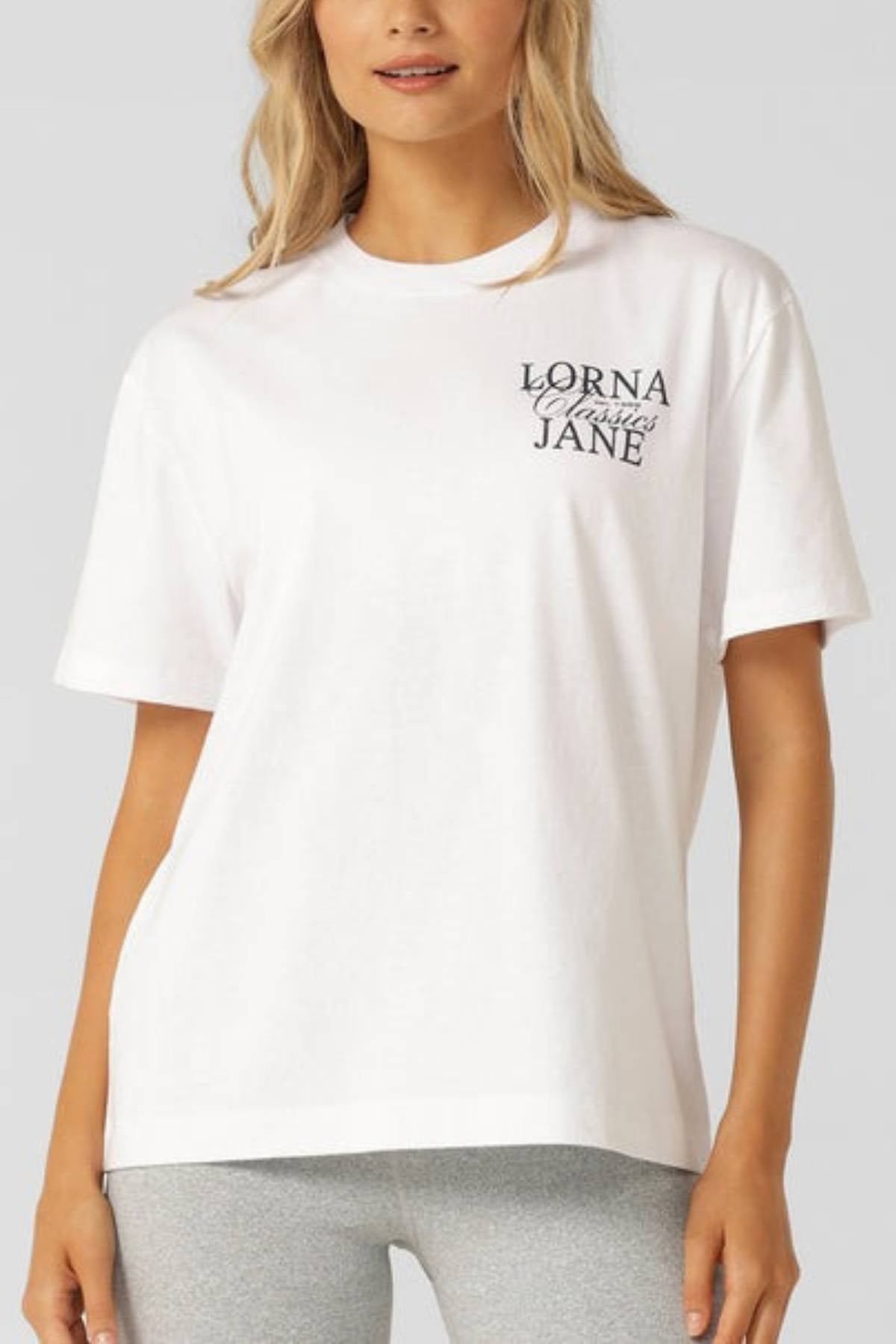 A woman wears the Lorna Jane Sprint Classic Fit T-Shirt in soft cotton jersey with "Lorna Jane" in black on the left chest, paired with light gray leggings. The background is plain and neutral.